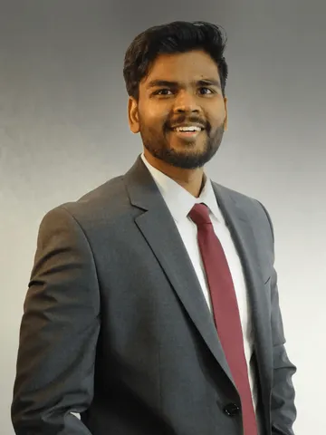 Aditya Kumar, smiling, wearing a grey suit and dark red tie against a grey studio background.