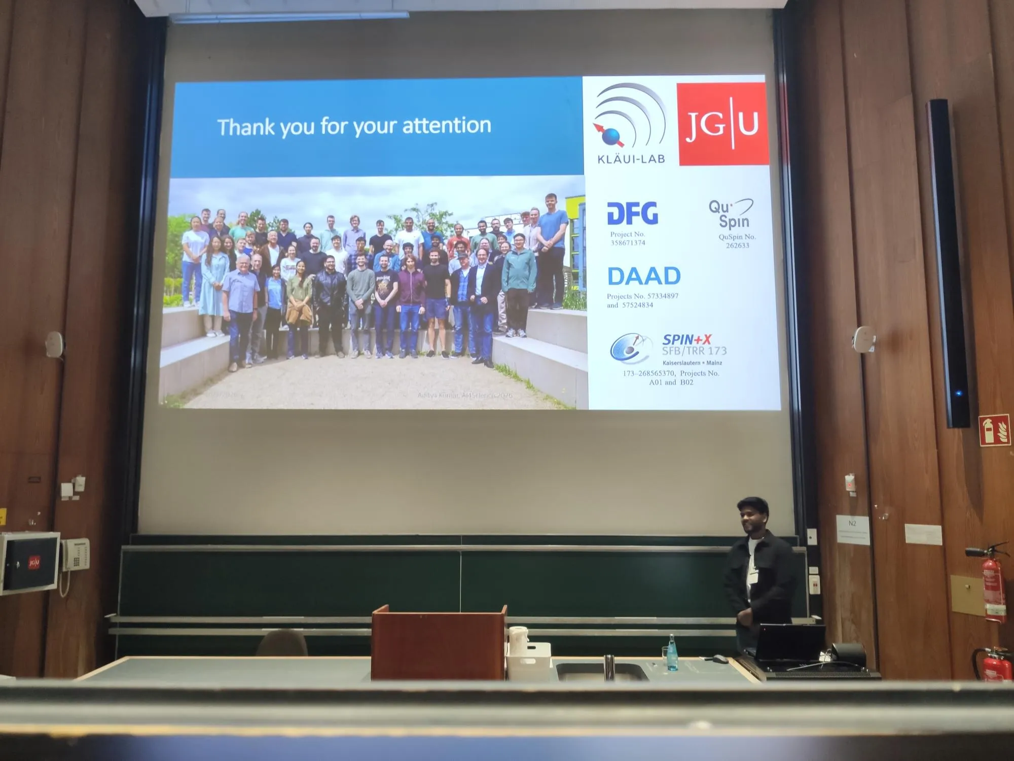 Aditya Kumar standing beside the lectern in a wood-panelled lecture hall at the end of his AI4Sci 2026 talk, below a projected closing slide reading Thank you for your attention, with a Kläui Lab group photograph and the logos of the Kläui Lab, JGU and the funding bodies.